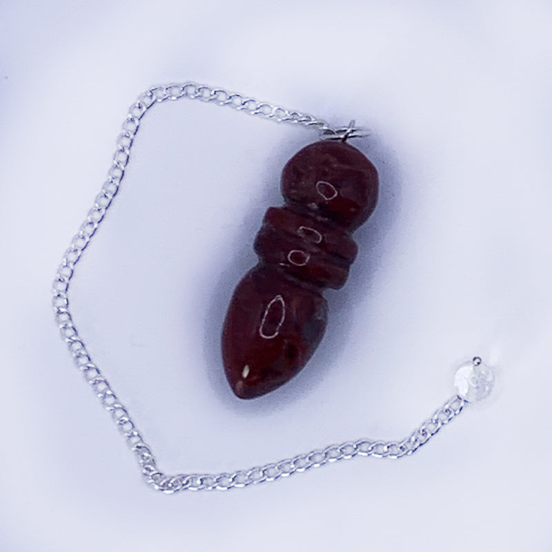 Pendule Egyptien en Jaspe Rouge suspendue à une chaîne argentée, forme sculptée et polie avec trois segments visibles.